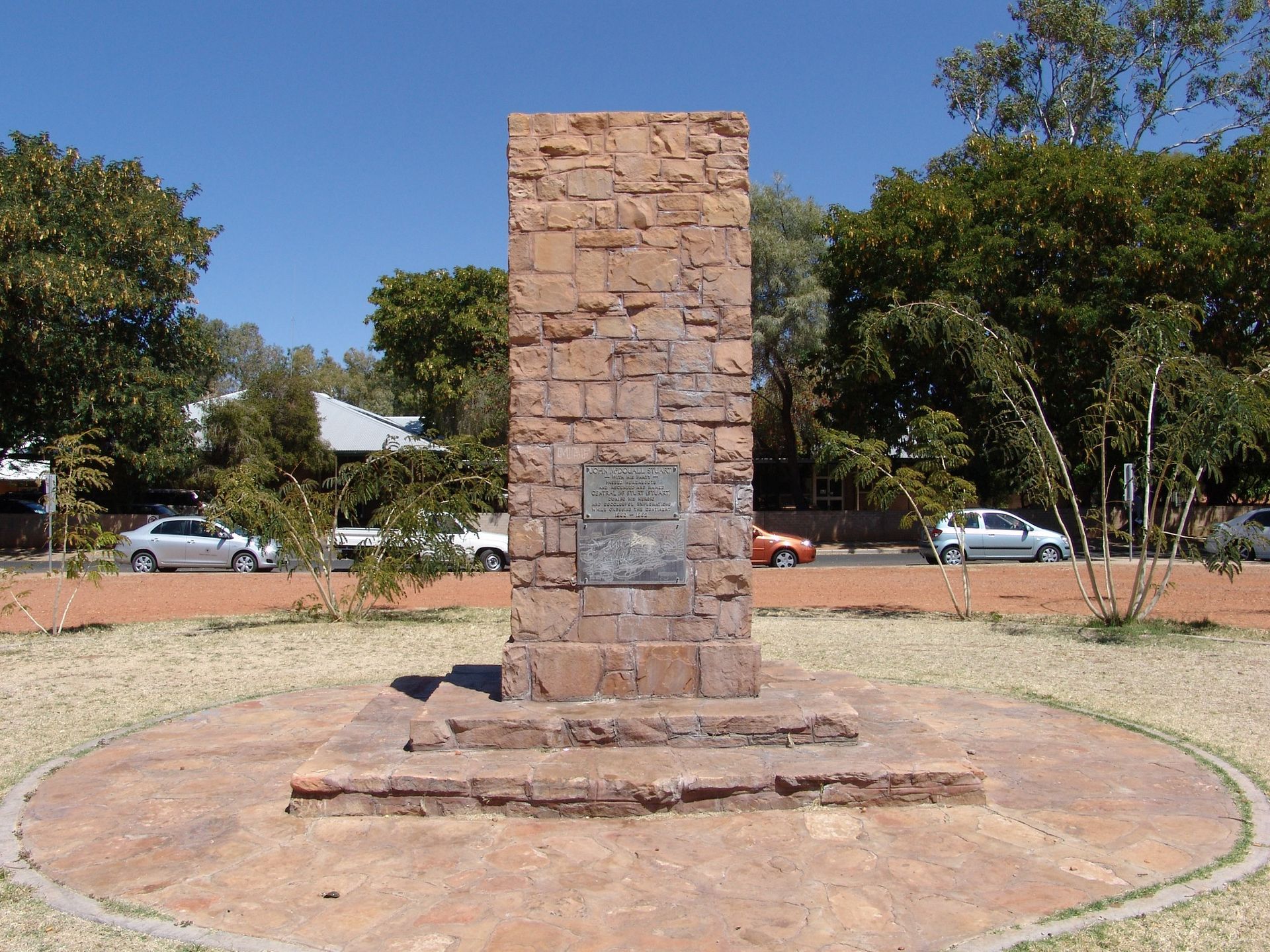Monument made of stone blocks in a circular plaza. Cars and trees in the background.