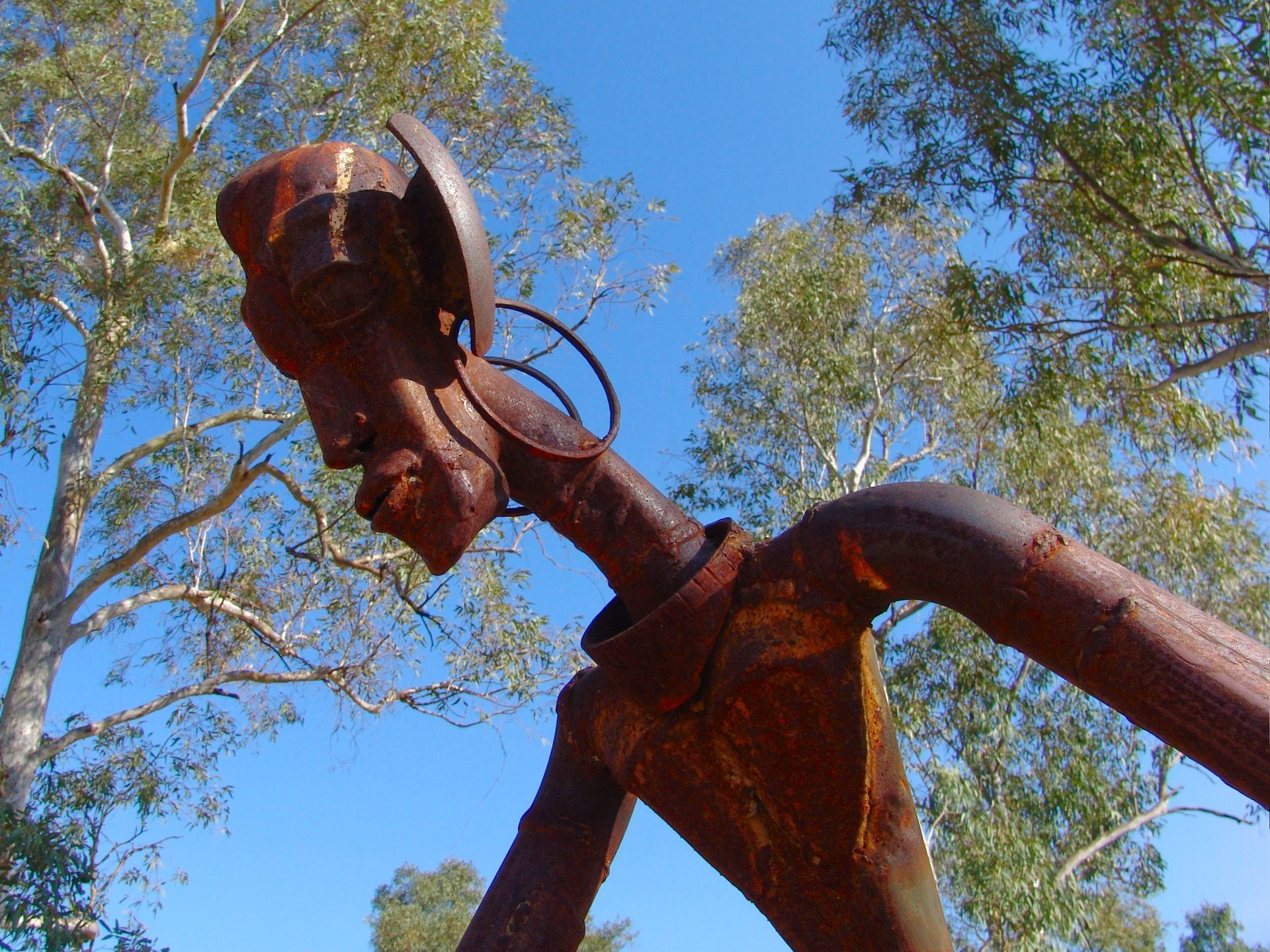 Rust-colored metal sculpture of a person, angled from below, set against a blue sky with trees.