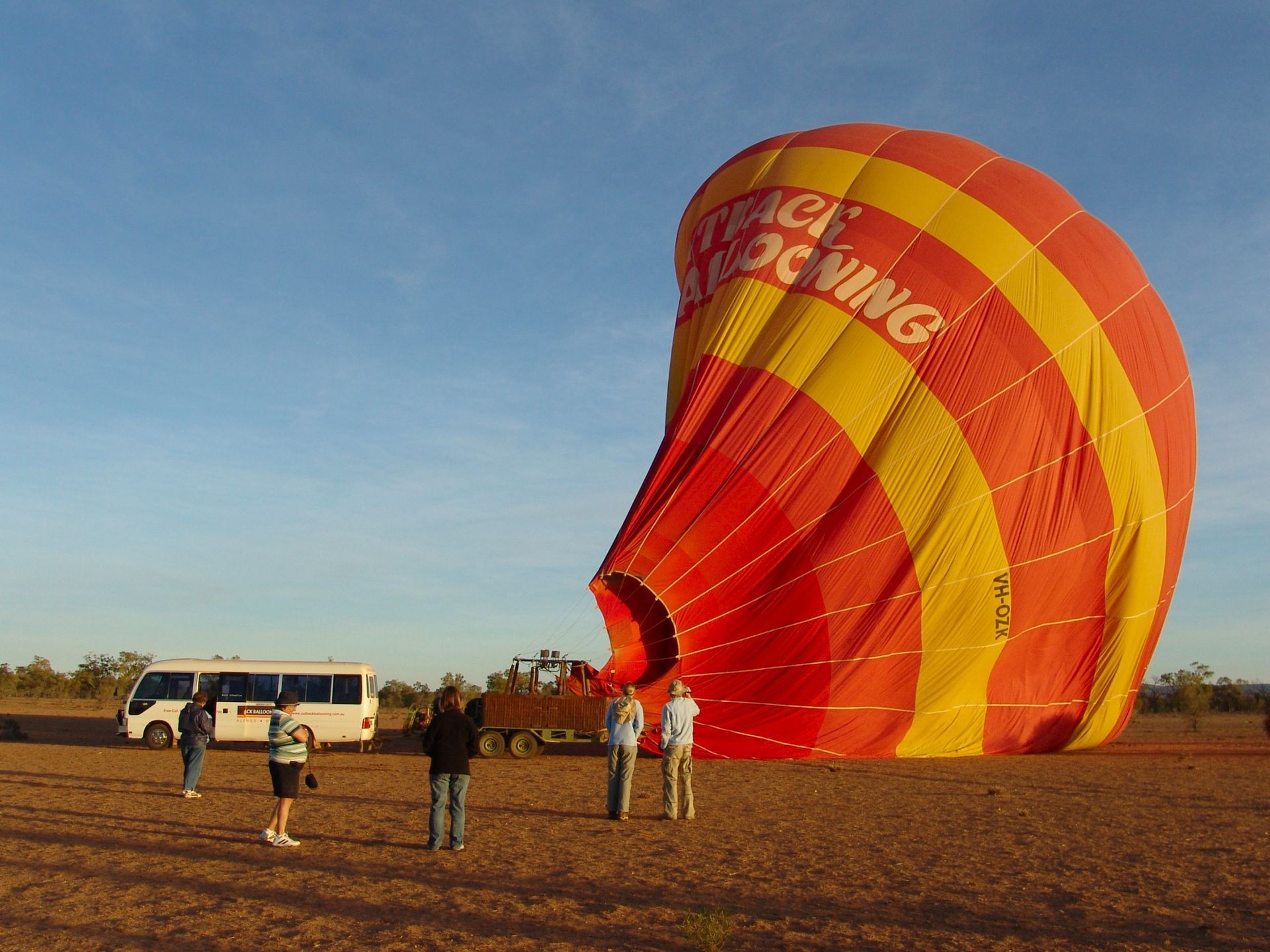 Hot air balloon on the ground with red and yellow stripes. People watch near a bus and truck on a field.