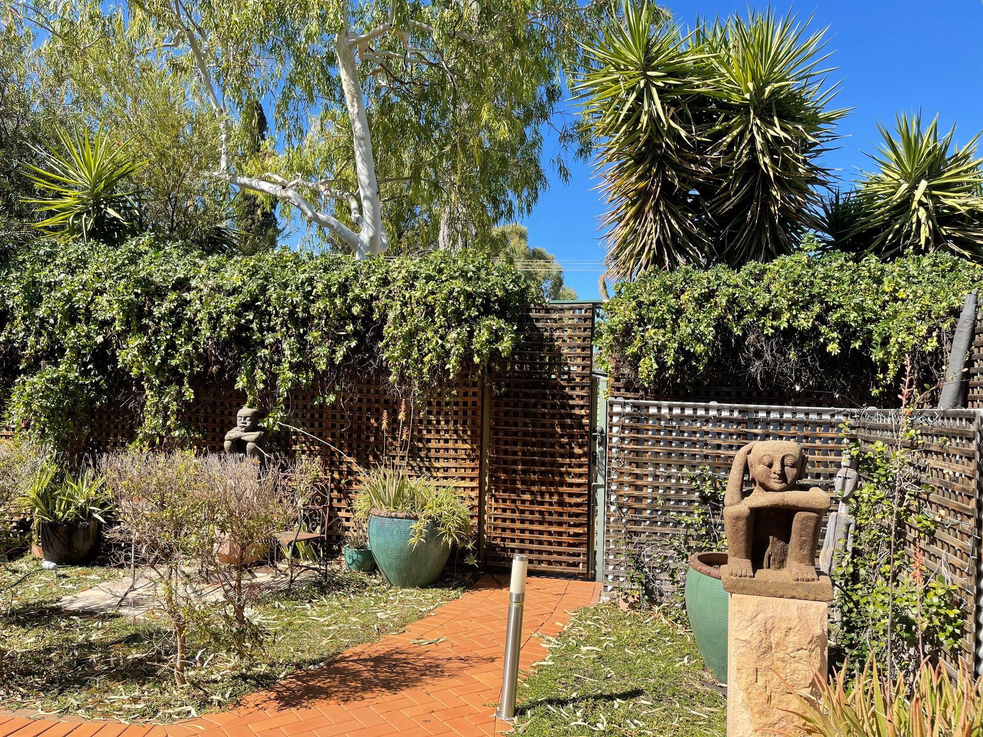 Lush garden with a red brick path, wooden lattice fence, and a stone statue.