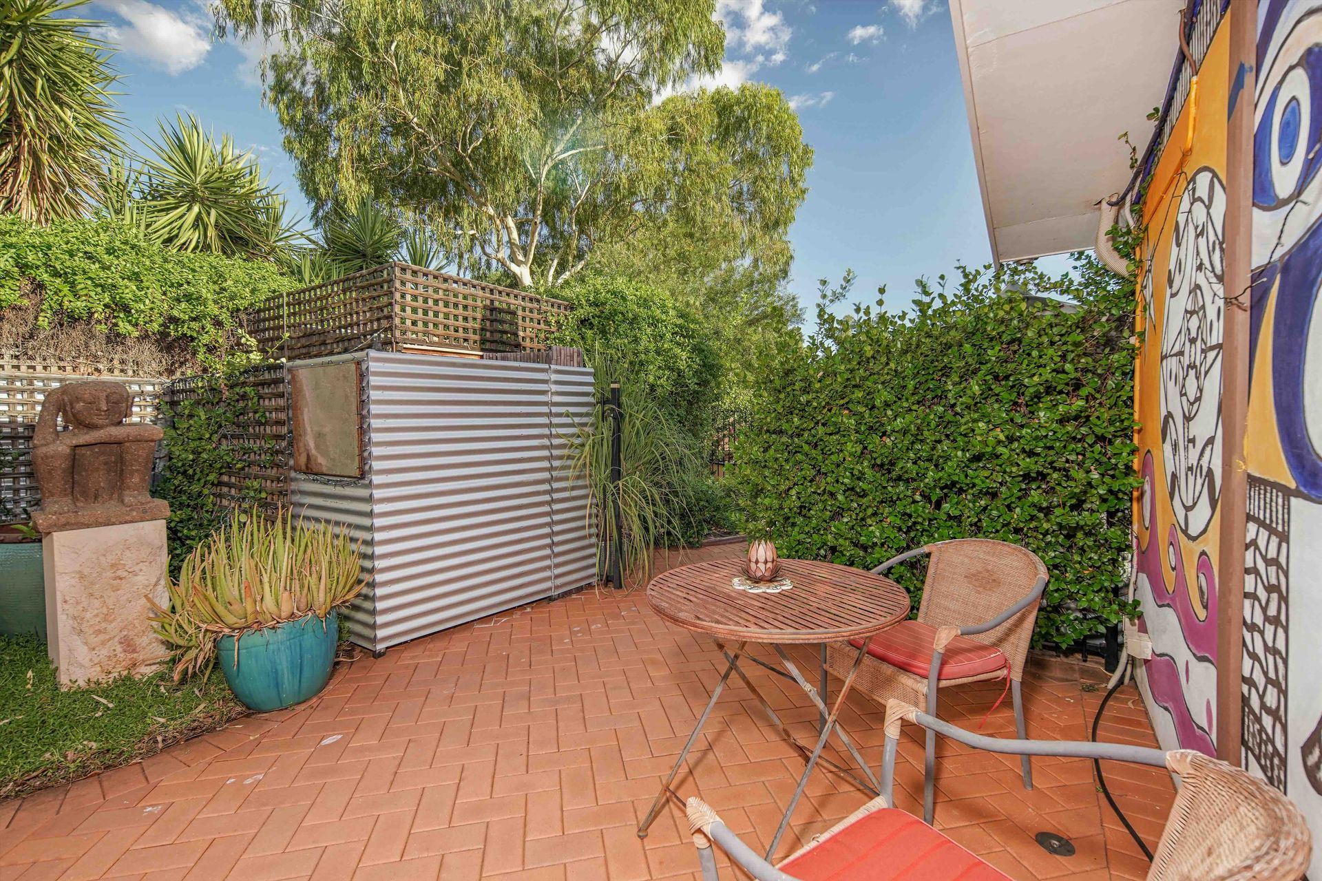 Brick patio with metal table and chairs, surrounded by greenery and art on the wall.