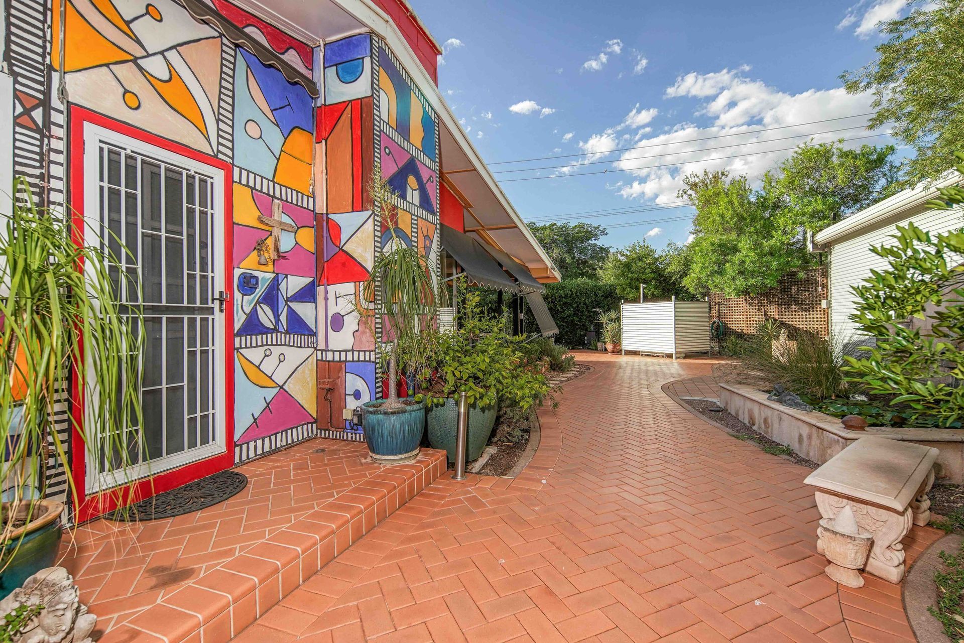 Colorful mosaic-covered building with a red-tiled patio, potted plants, and a walkway leading to a backyard.