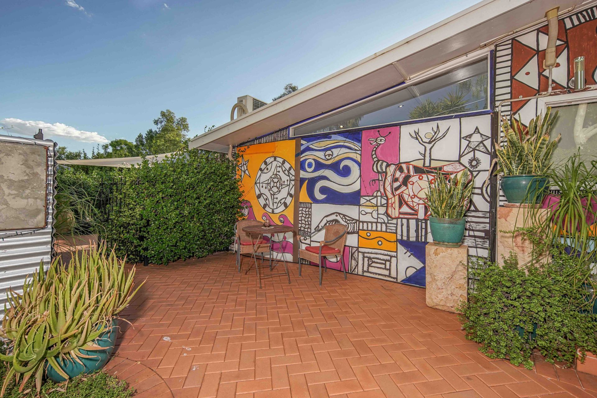 A colorful patio area with painted walls, plants, brick flooring, and a small table with chairs.