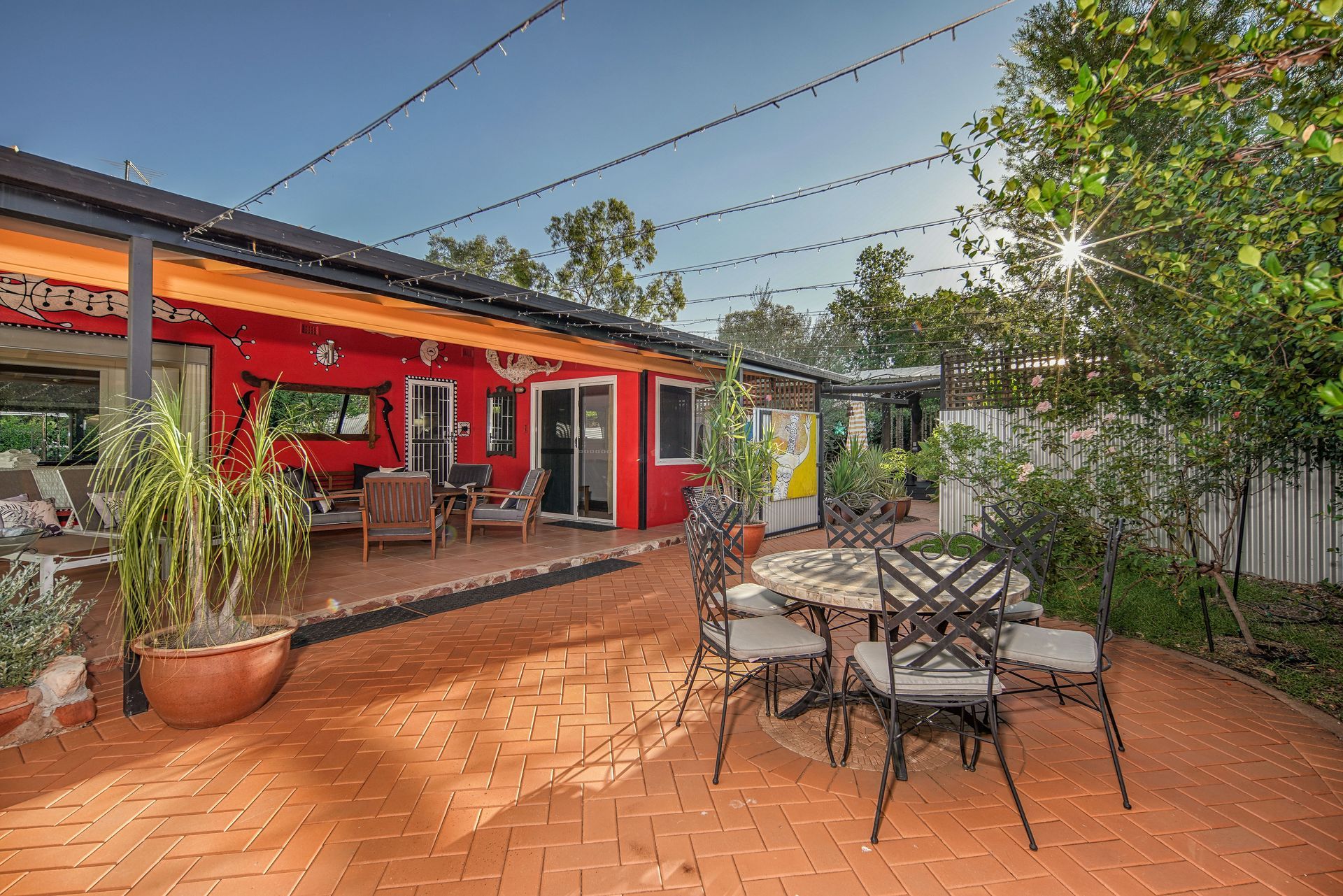 Red-walled patio with outdoor seating, brick pavers, and string lights; trees and bright sunlight in the background.