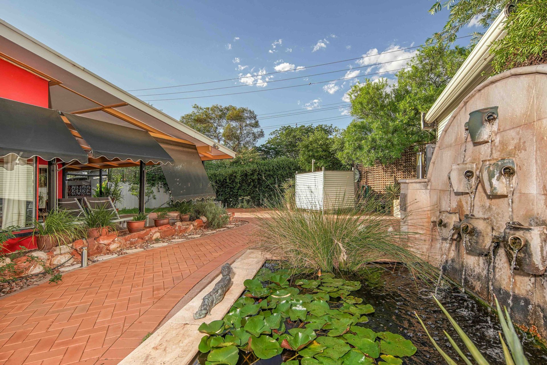 Brick pathway leads to building with awning, next to a pond and fountain. Blue sky with clouds.