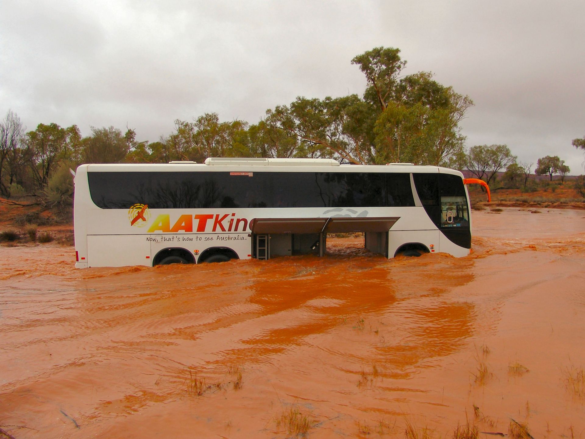 A white AAT Kings bus stuck in muddy, flooded terrain. Brown water surrounds the vehicle in a desert landscape.