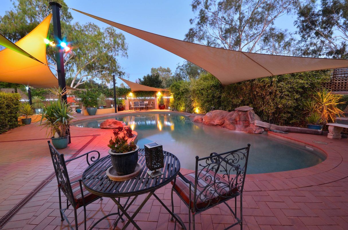 Swimming pool with shade sails, patio furniture, and surrounding greenery.