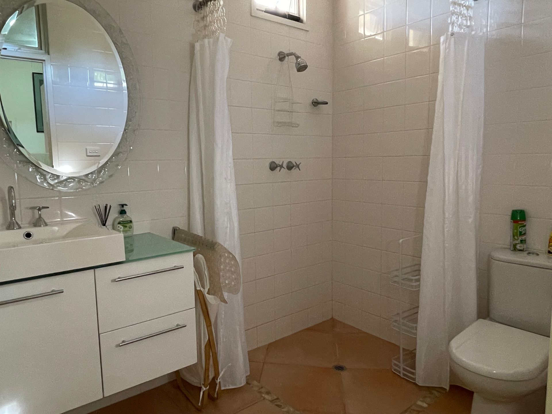 Bathroom with a white vanity, oval mirror, shower, and toilet. Beige tiles and white curtains.