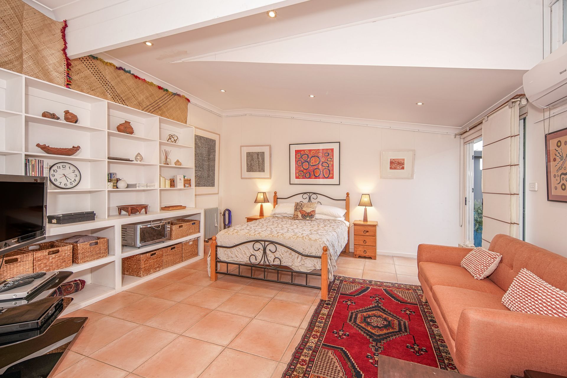 Bedroom with bed, sofa, shelves filled with items, terracotta floor.