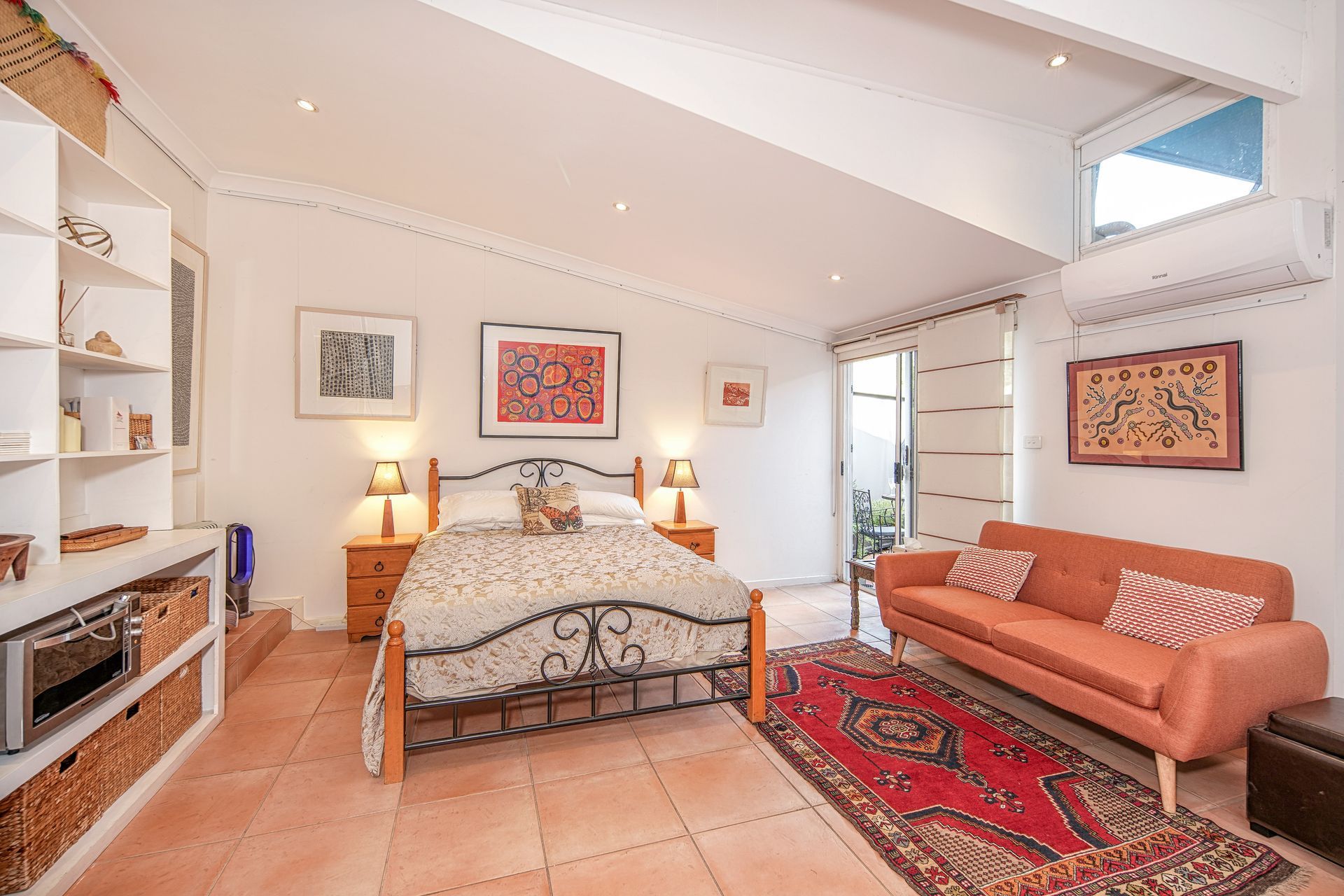 Bedroom with bed, orange couch, rug, art, and open door to an outdoor area.
