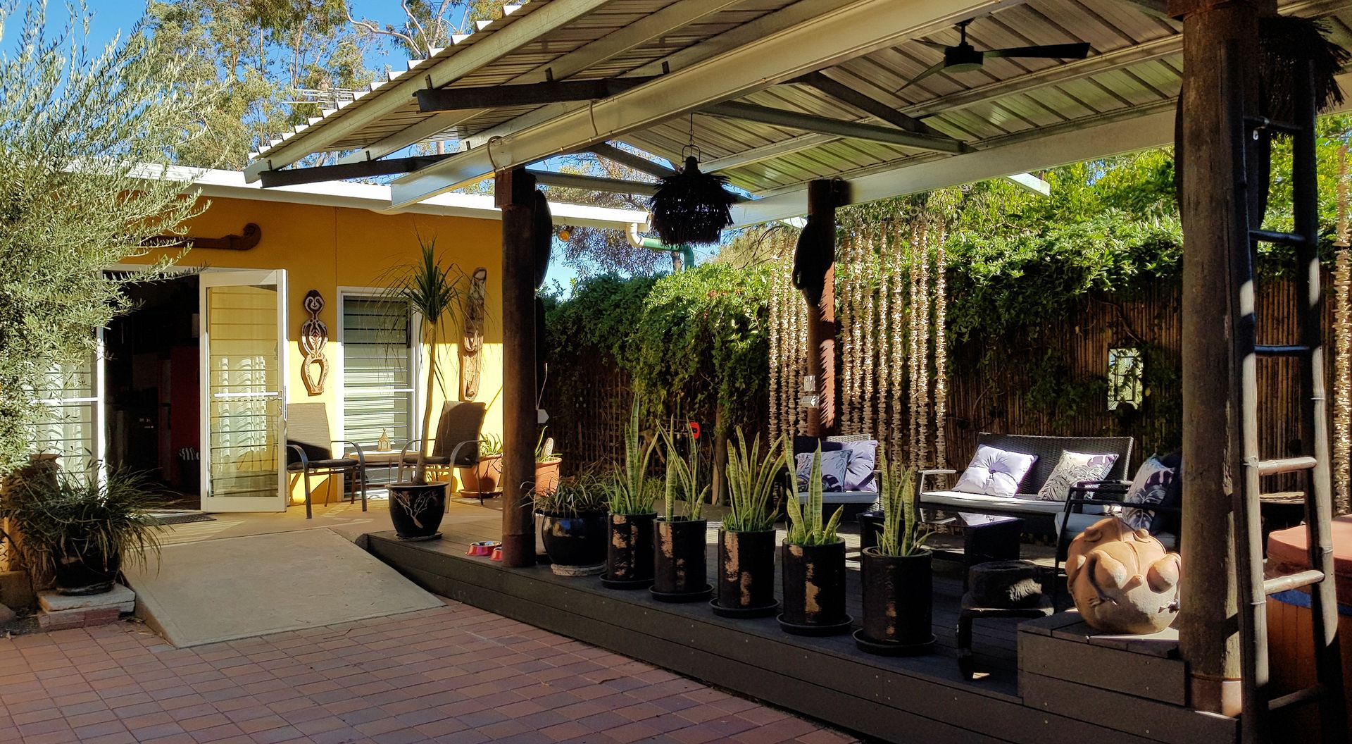 Patio with covered seating, potted plants, and ramp leading to a yellow building with an open door.