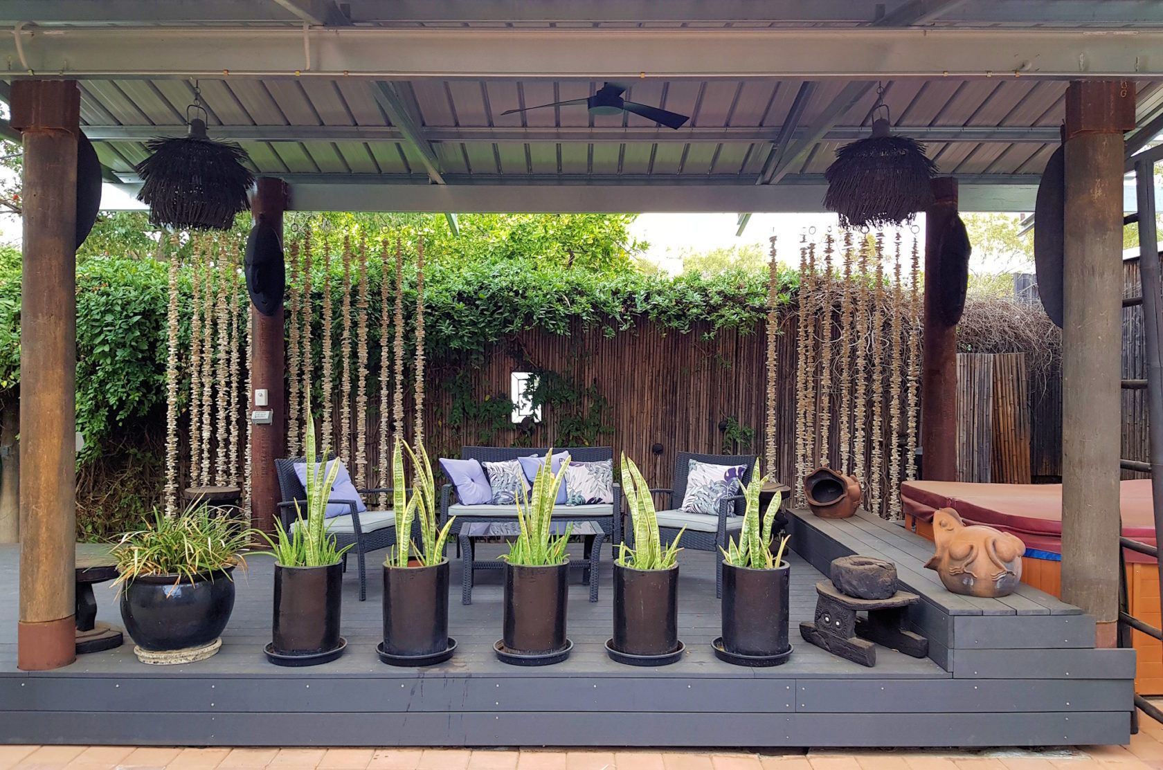 Outdoor seating area with grey furniture, potted plants, and bamboo backdrop under a canopy.
