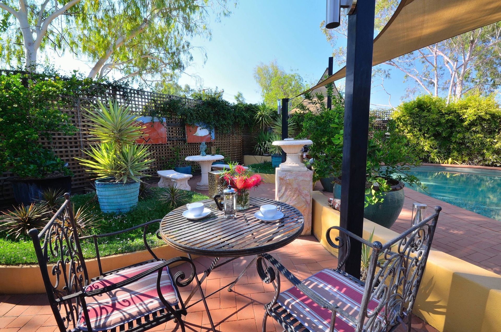 Outdoor patio with table set for two, near a pool, with plants and shade.