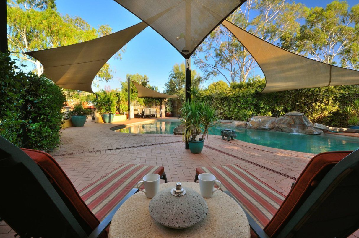 Lounge chairs with coffee overlooking a pool, shaded by a tan canopy. Lush greenery surrounds the water.