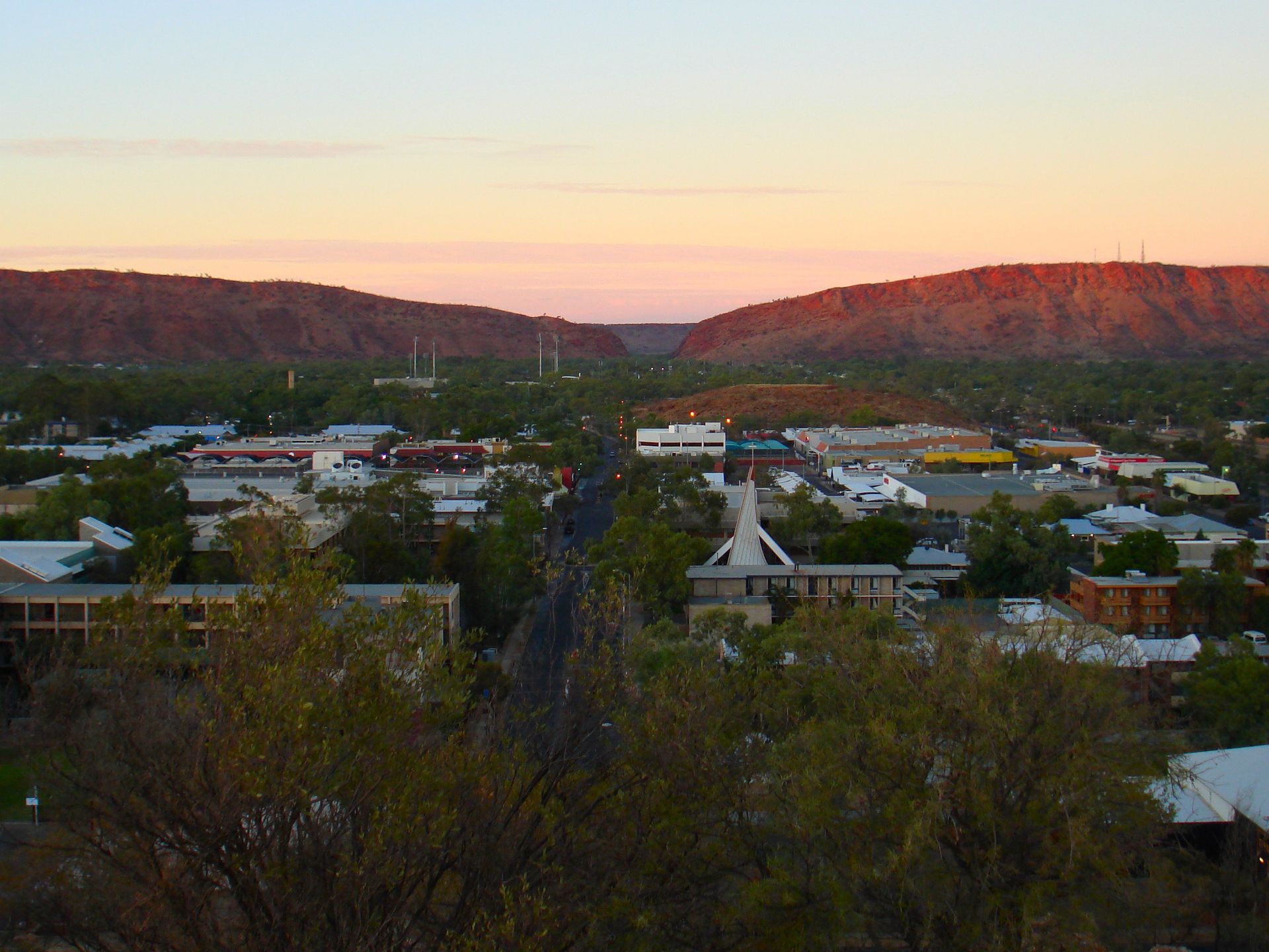 Town at sunset, with buildings, road, and mountains in the background under an orange and pink sky.