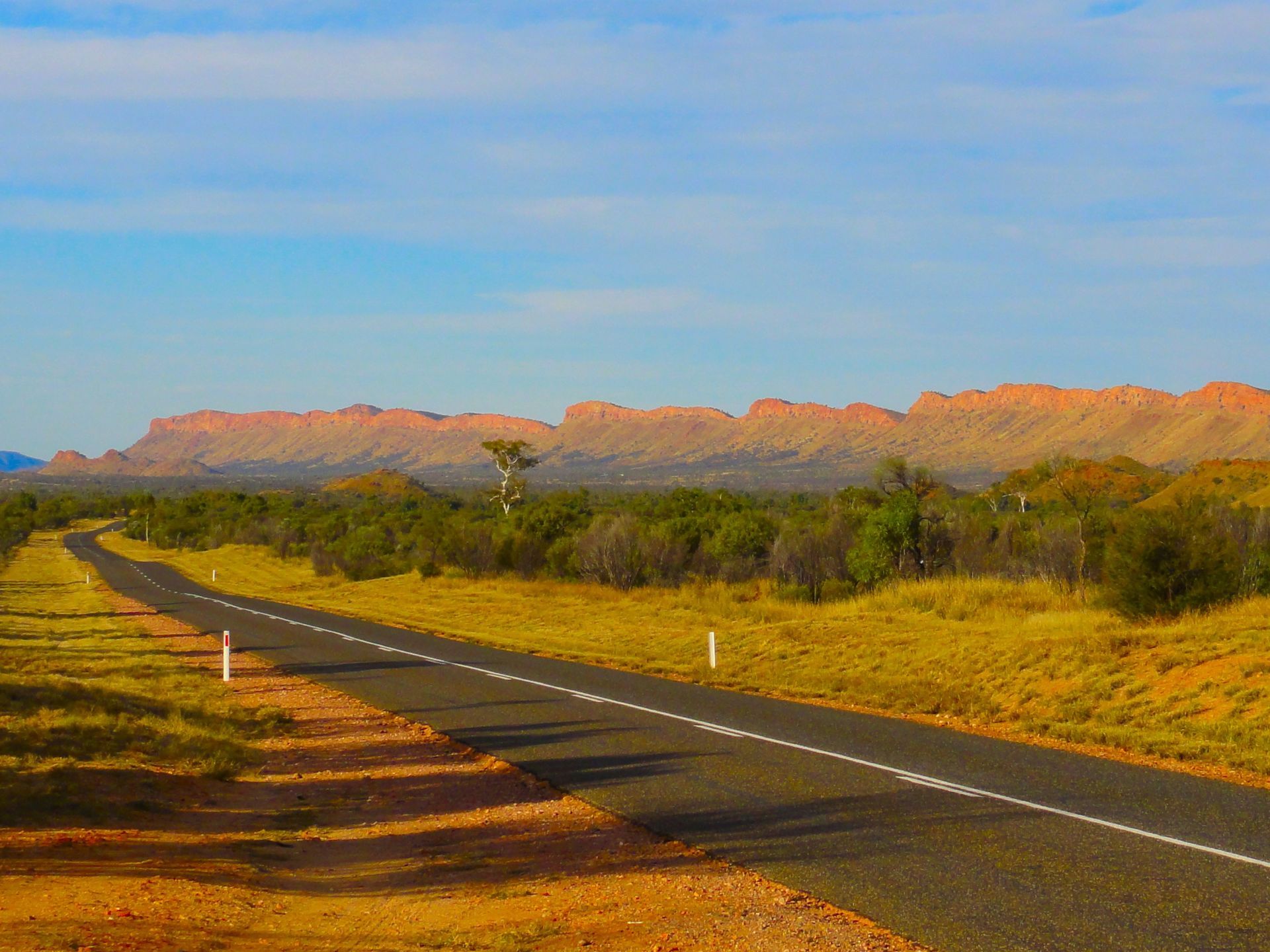 Road through arid landscape towards red rock formations under a blue sky.