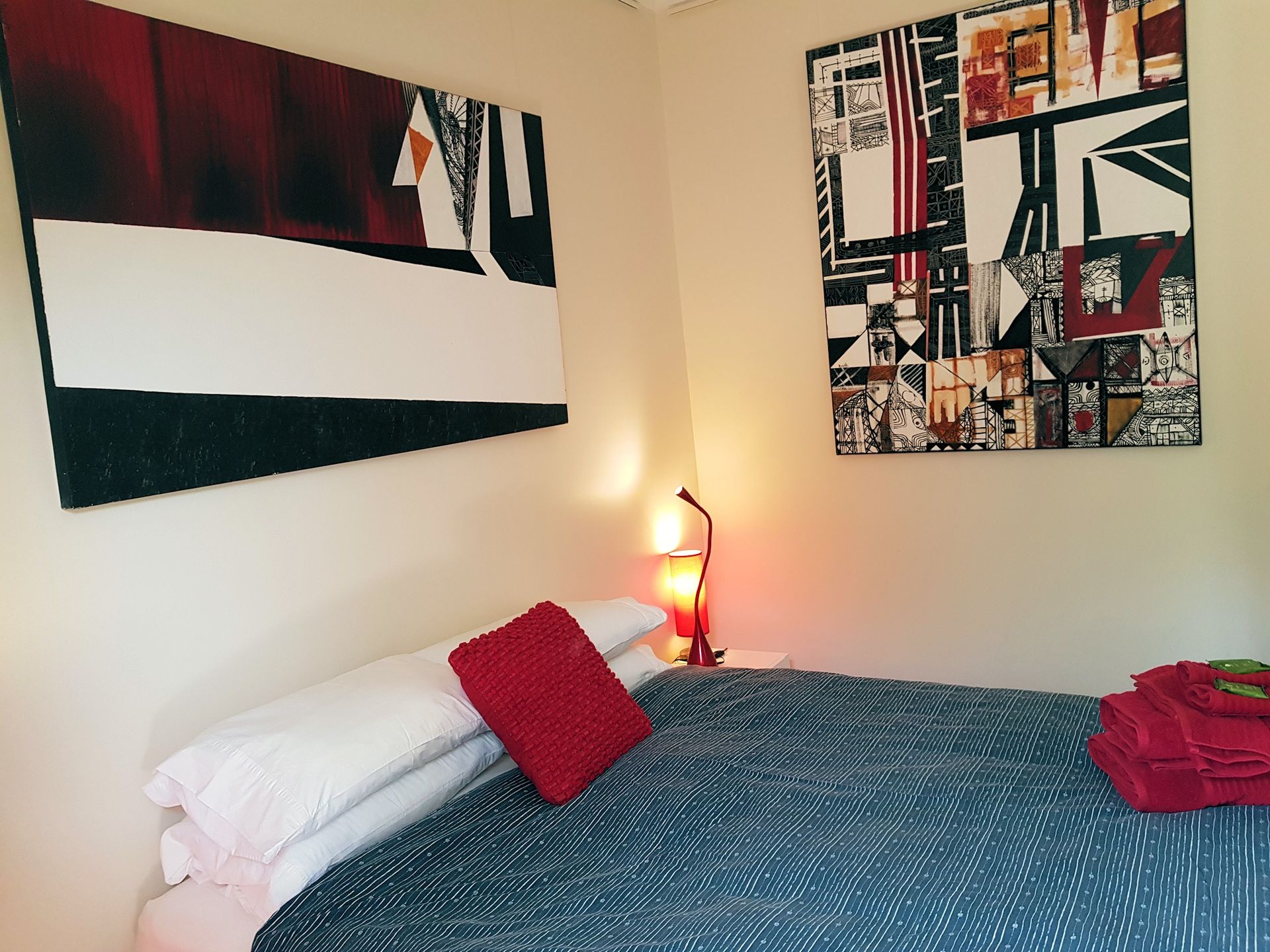 Bedroom with bed, art, and a bedside lamp. Red accents on pillows, artwork, and towels.
