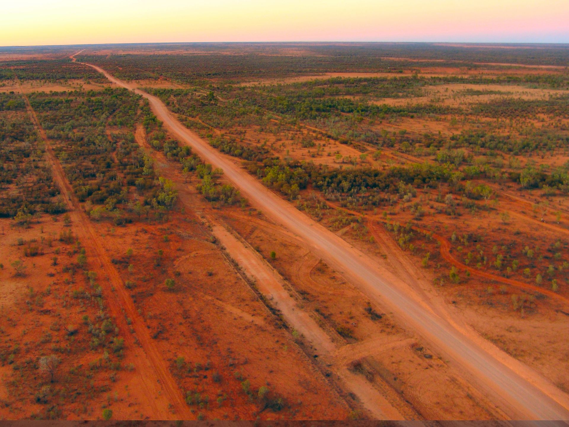 Dirt road through arid, reddish Australian outback landscape, sparse vegetation under a sunset sky.