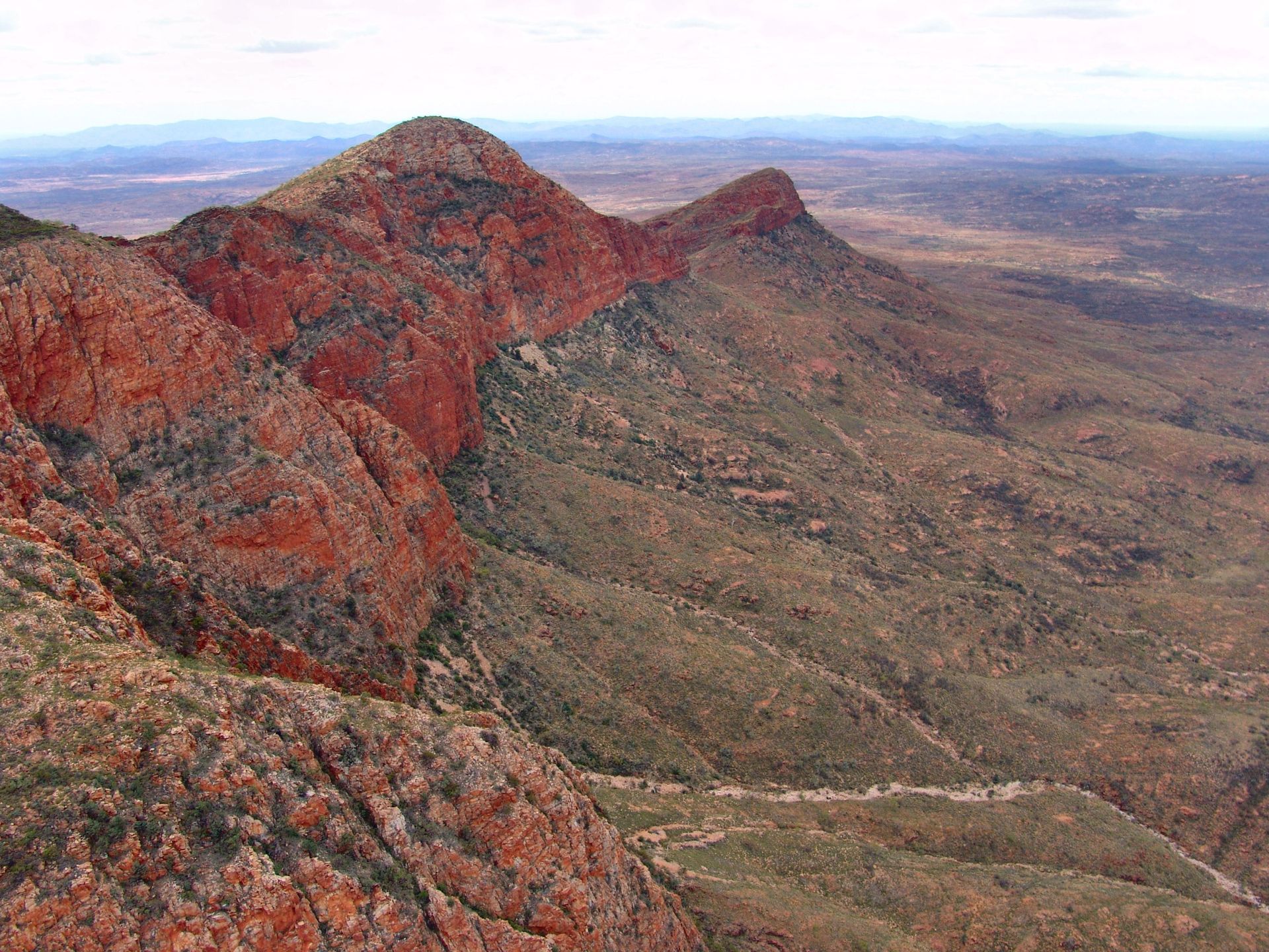 Red and green mountains in a dry, desert landscape under a hazy sky.
