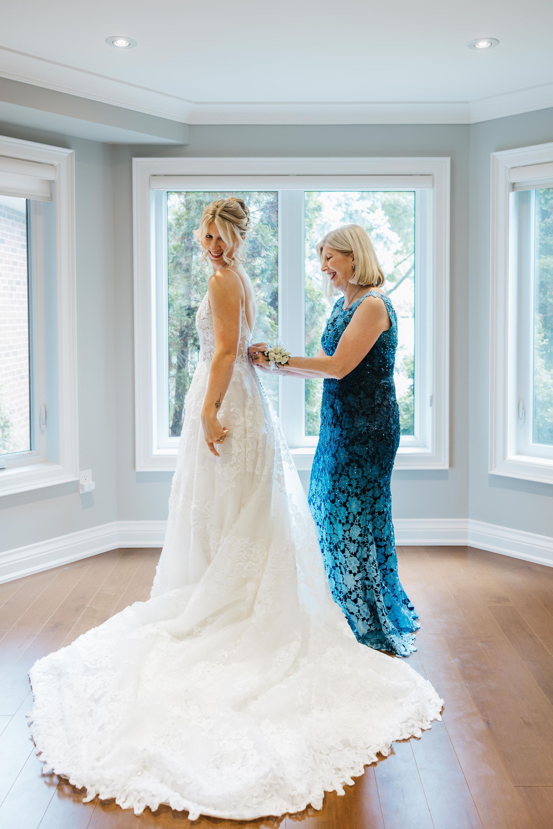 Bride in white gown being adjusted by a woman in a blue dress in a bright room