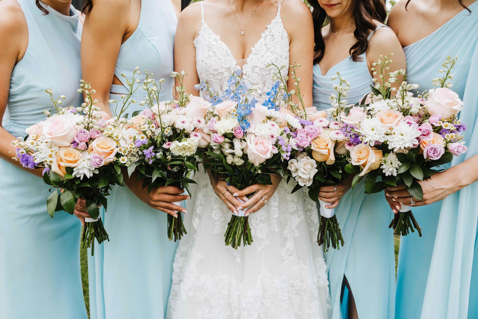 Bridesmaids in light blue dresses holding pastel flower bouquets beside a bride in white lace dress