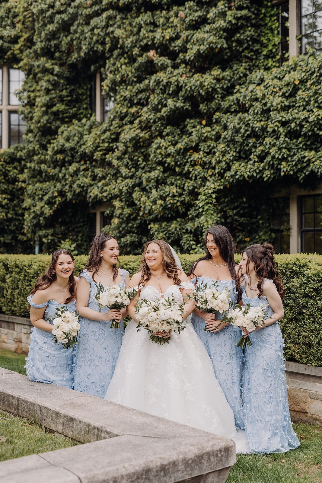 Five women in blue dresses and a bride in a white gown holding bouquets in front of ivy-covered walls.