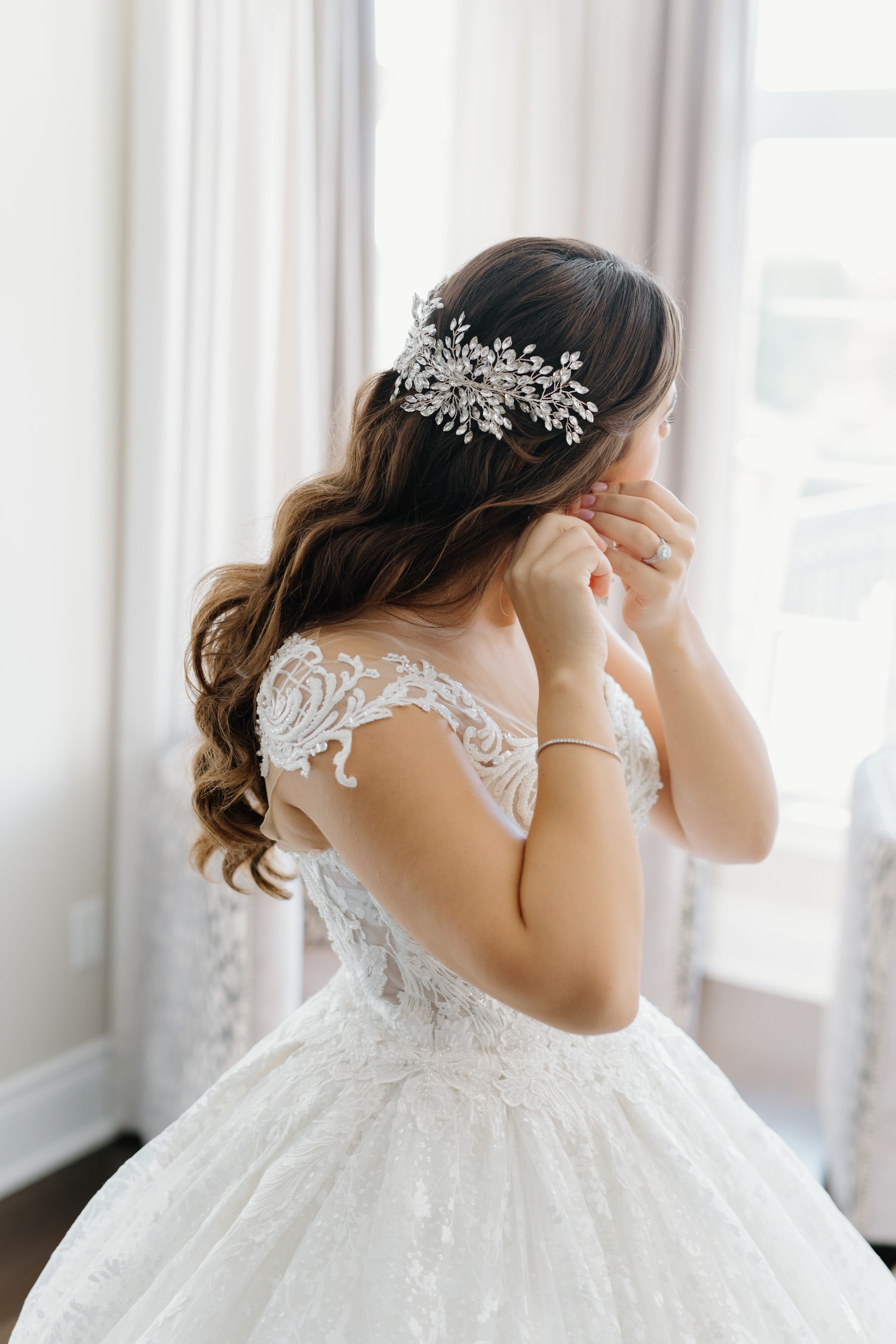 Bride in a white lace gown adjusting her earring by a bright window