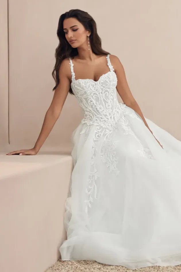 Woman in a white lace wedding dress seated against a beige backdrop.