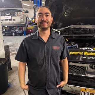 Mechanic in a black uniform standing in an auto repair shop with cars on lifts behind him | Accurate Automotive Northglenn