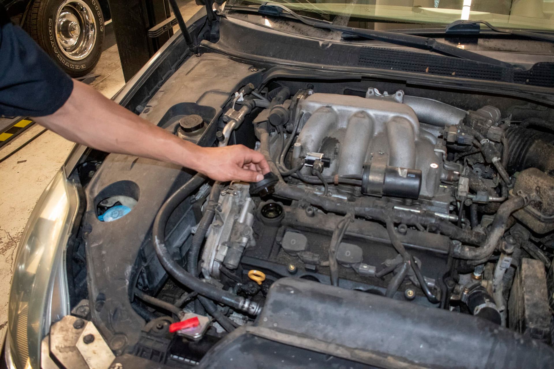 Person inspecting a car engine bay with a hand near hoses and wiring under the hood | Accurate Automotive Northglenn