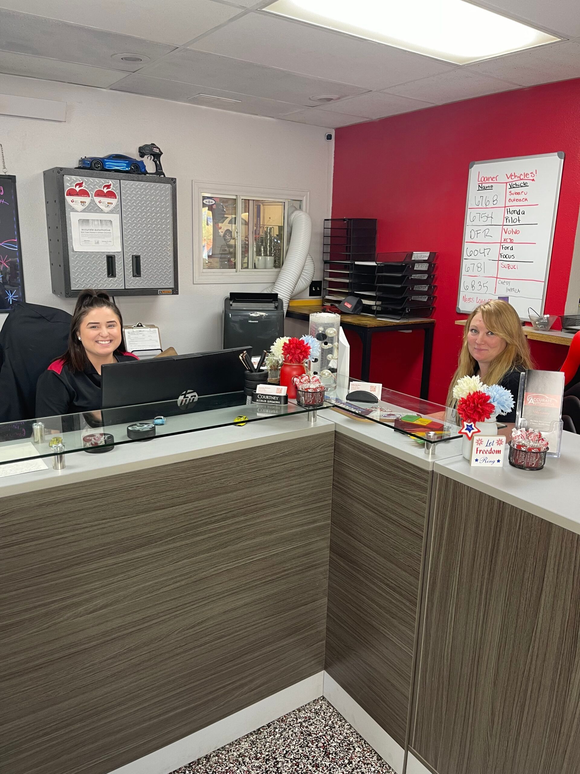 Reception desk with two staff members, red accent wall, and office equipment in the background | Accurate Automotive Northglenn