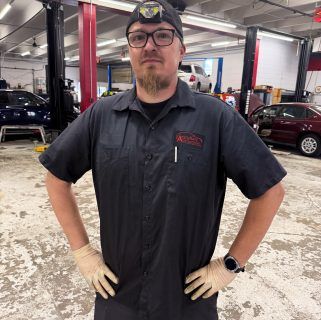 Mechanic in dark work shirt and cap standing in auto shop with cars and red lifts, hands on hips. | Accurate Automotive