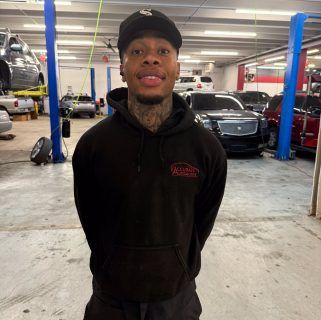Man standing in a car repair shop, wearing a black hoodie and cap, with vehicles and lifts in the background | Accurate Automotive Northglenn