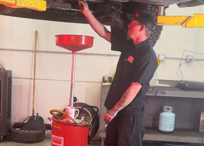 Mechanic inspecting a car on a lift, using a red oil drain pan in an auto shop. | Accurate Automotive Northglenn