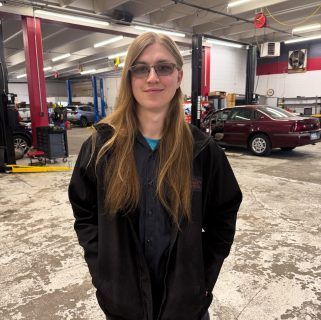 Person standing in an auto repair garage, wearing glasses and a black jacket, with a red car in the background. | Accurate Automotive Northglenn