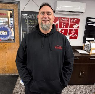 Man in a black hoodie smiles, standing indoors in an office or shop with signs and cabinets behind him | Accurate Automotive