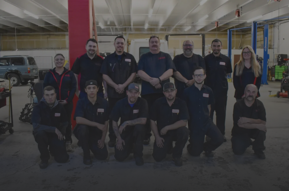 Group of workers in black uniforms posing in a workshop, with yellow equipment and a red vehicle in the background. | Accurate Automotive Northglenn