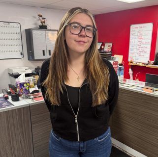 Person standing in an office, wearing glasses and a black top, with a red accent wall and desk behind them | Accurate Automotive
