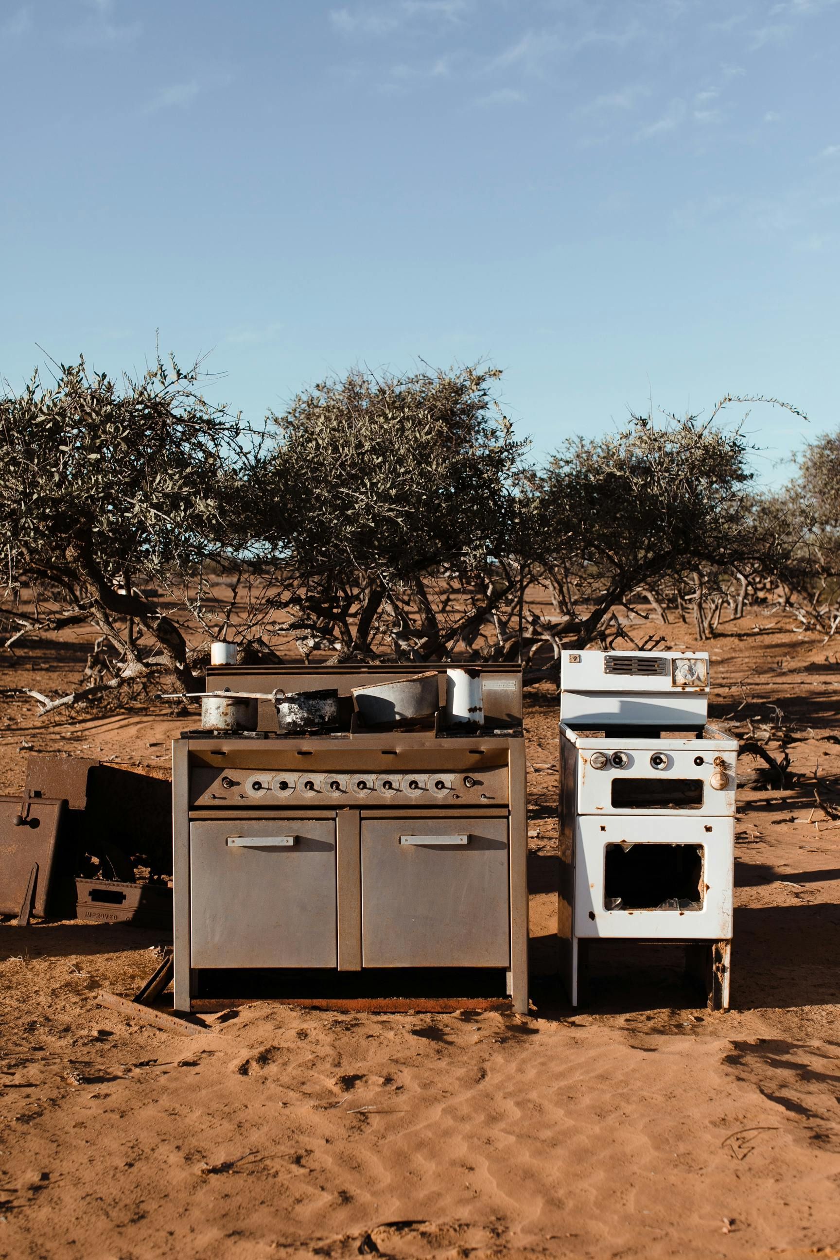 Old kitchen appliances abandoned in a dry, sandy landscape with trees under a blue sky.