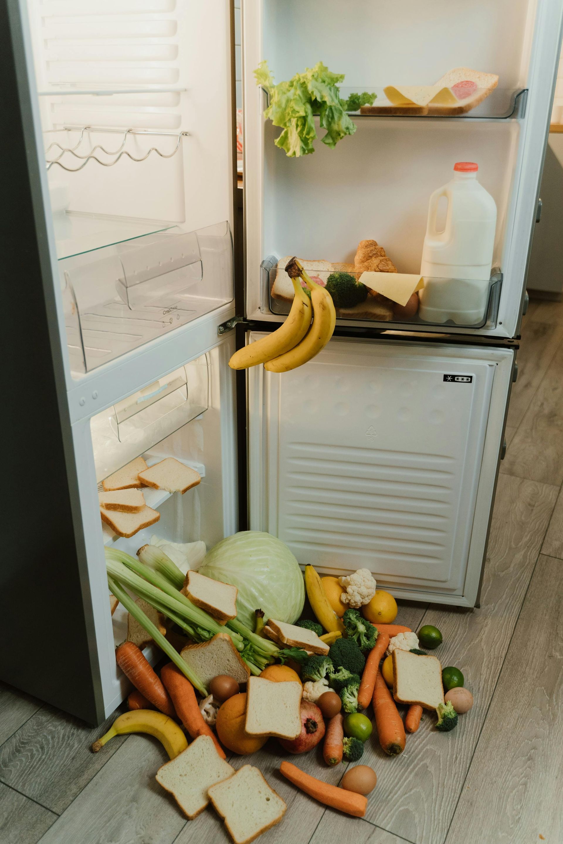 Refrigerator with door open, food spilling out onto floor: bread, bananas, vegetables, and milk.