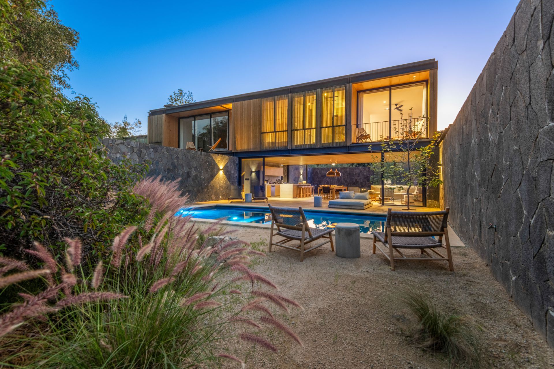 Casa moderna con ventanas iluminadas, piscina y terraza con asientos al atardecer.