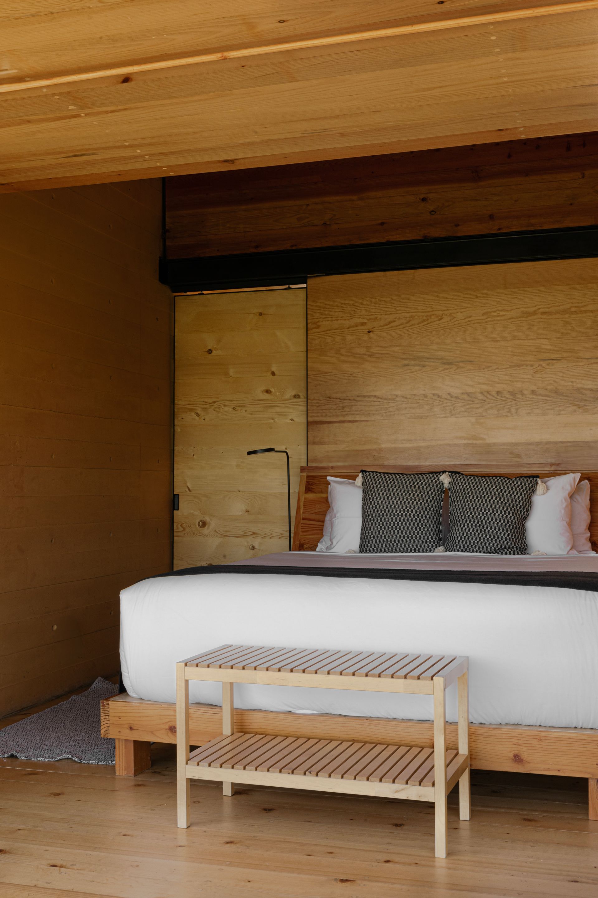 Dormitorio minimalista de madera con cama blanca, banco tejido y paneles de pared rústicos.