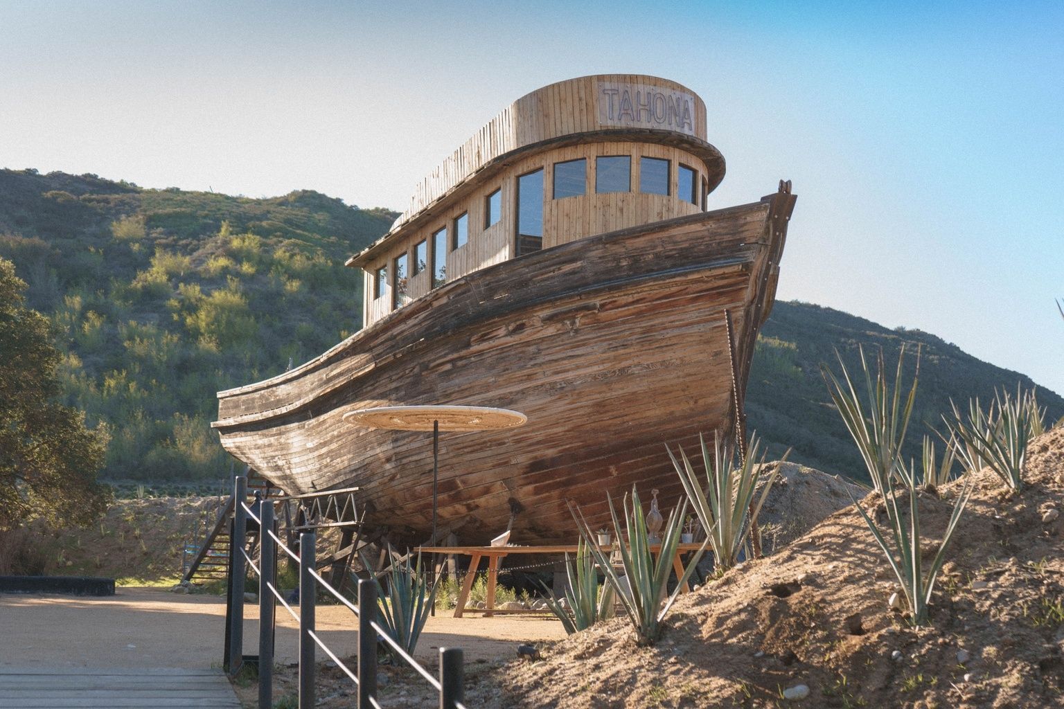 Edificio de madera con forma de barco sobre pilotes junto a plantas desérticas y colinas bajo un cielo despejado.