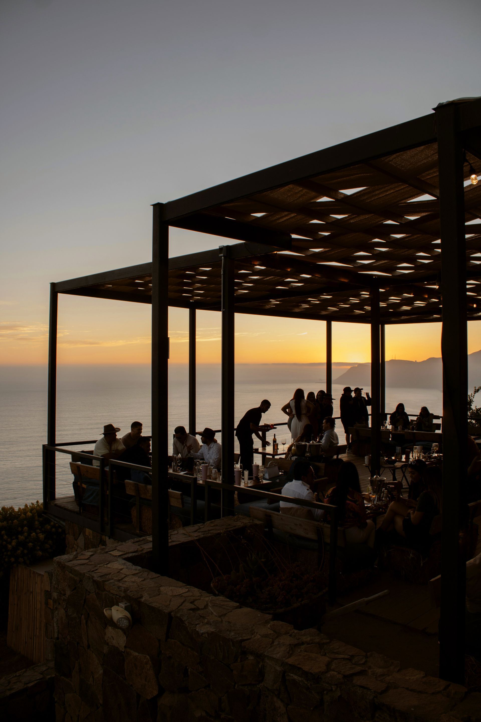 Gente cenando en una terraza junto al mar al atardecer, recortadas contra una pérgola oscura.