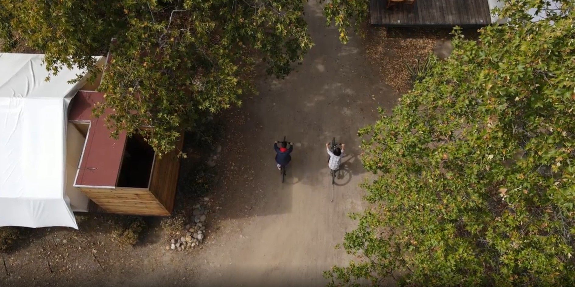 Dos personas caminando por un sendero de tierra junto a tiendas de campaña y árboles, vistas desde arriba.