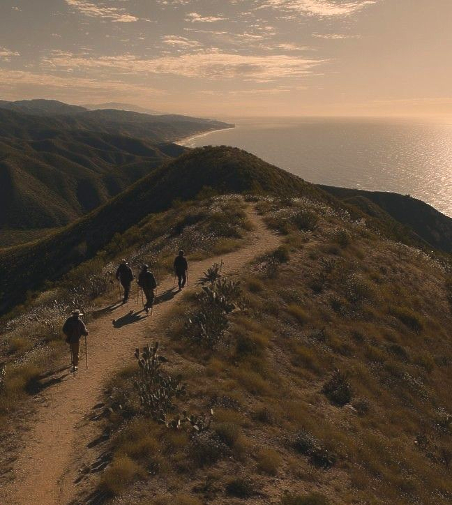 Senderistas caminan por una cresta costera iluminada por el sol sobre el océano al atardecer.