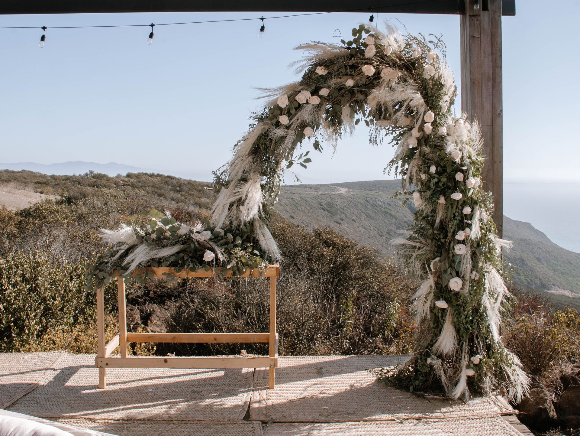 Arco floral para bodas junto a un banco de madera con vistas a una ladera costera bañada por el sol.