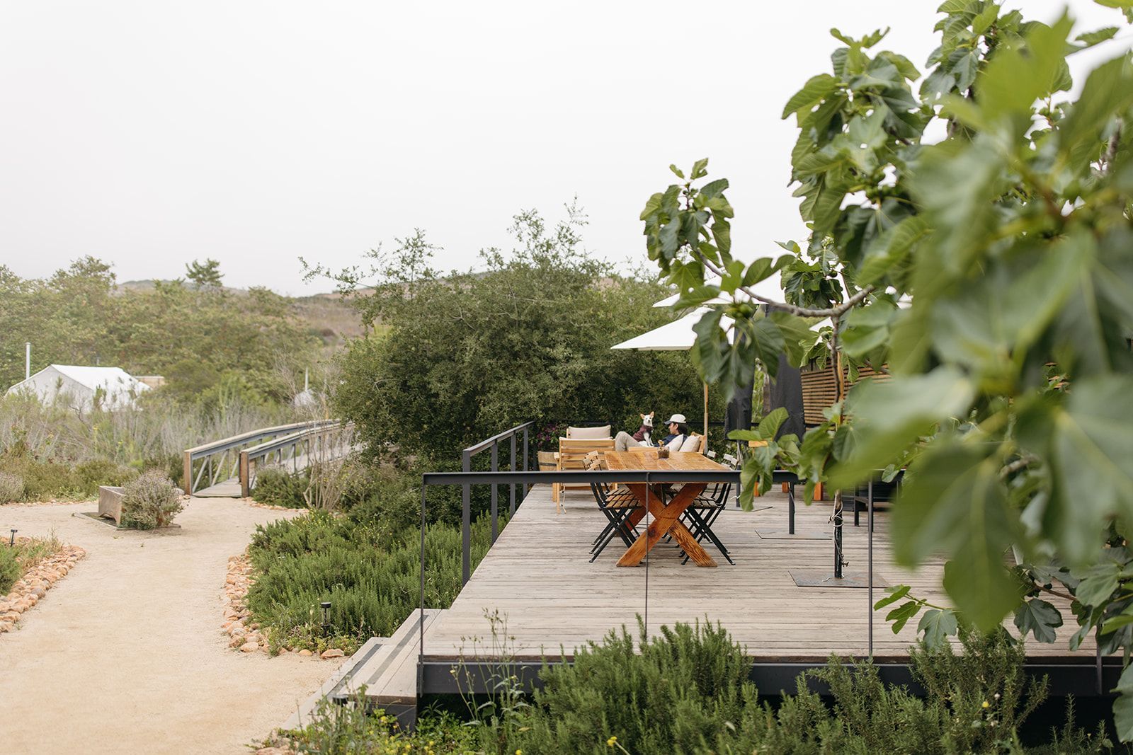Patio ajardinado con terraza de madera, mesa y sillas naranjas y sendero arenoso junto a la vegetación.