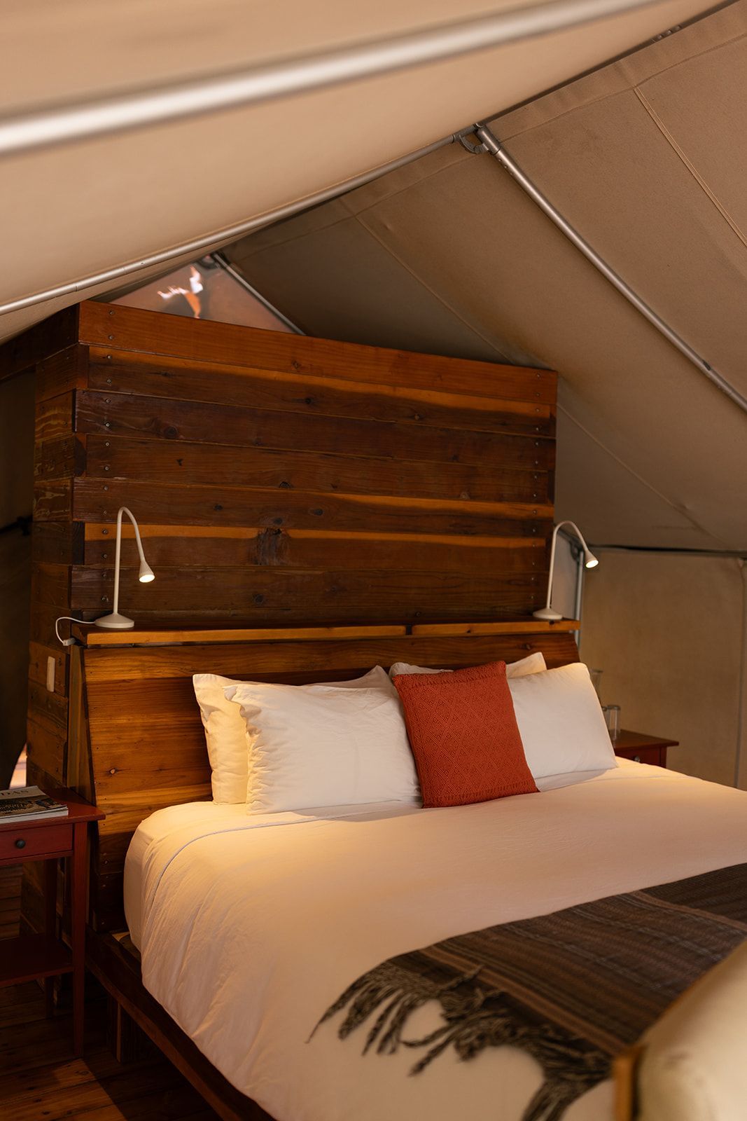 Cama acogedora con ropa de cama blanca, almohadas naranjas y cabecero de madera dentro de una habitación con forma de tienda de campaña.