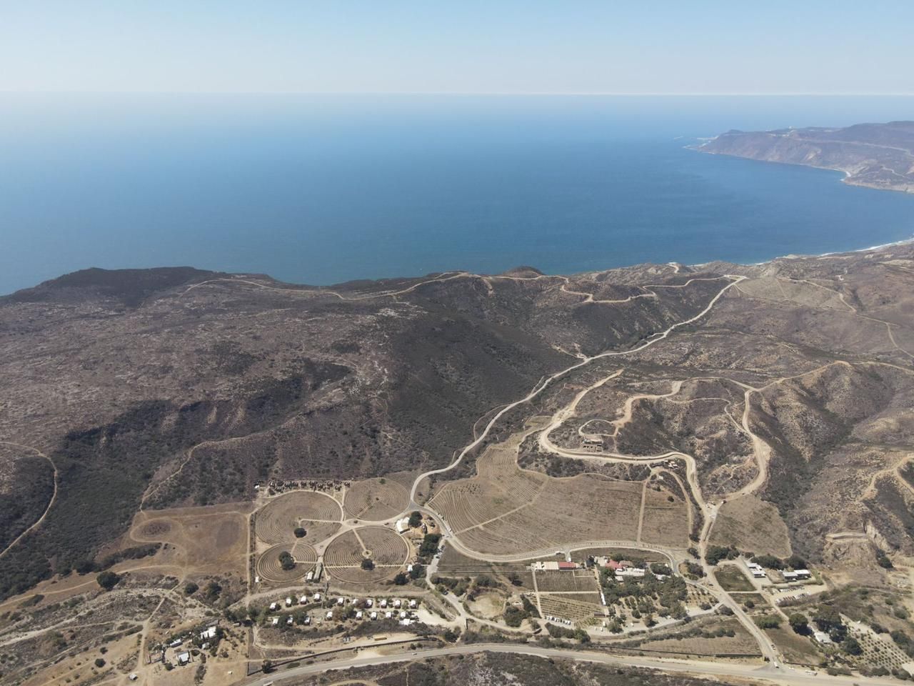 Vista aérea de un paisaje costero accidentado con carreteras sinuosas y el mar azul al fondo.